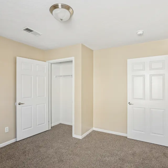 A vacant room featuring beige walls and a carpeted floor. There are two white doors, one leading to a closet and the other to another room or hallway. A ceiling light is visible.