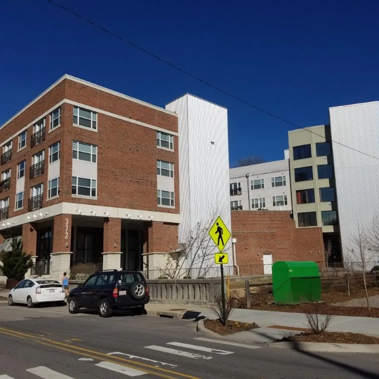 A modern building complex featuring a combination of brick and white panel architecture, with multiple stories and surrounding landscape elements. There is a crosswalk in the foreground and parked cars.