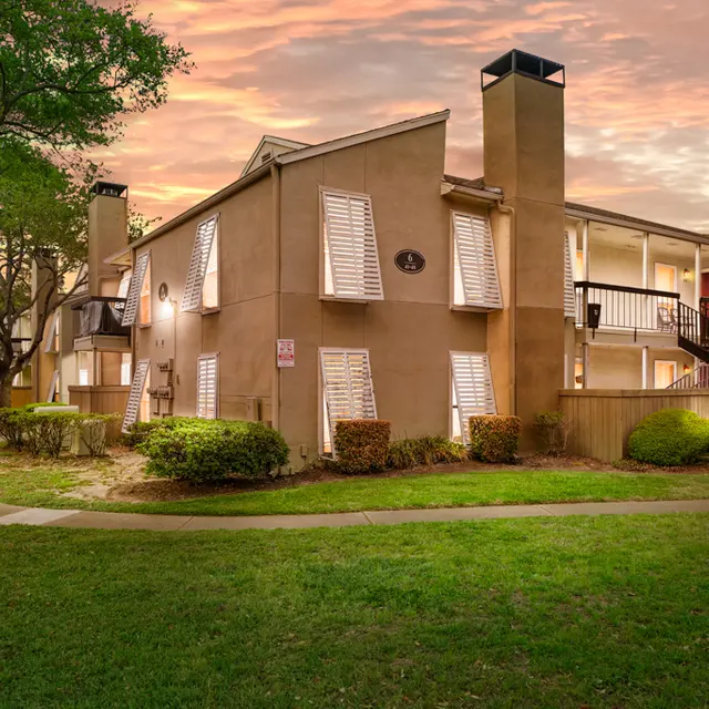 A view of an apartment complex at sunset with a well-manicured lawn, trees, and walkways in the foreground. The buildings feature cream-colored exteriors and balconies.