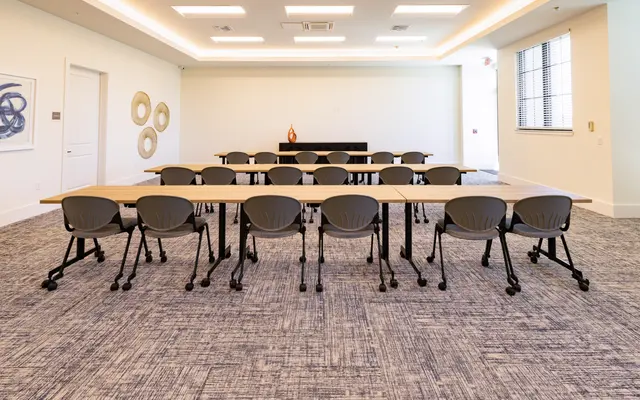 A modern meeting room featuring a long arrangement of tables and ergonomic chairs, with a minimalist design and neutral colors.