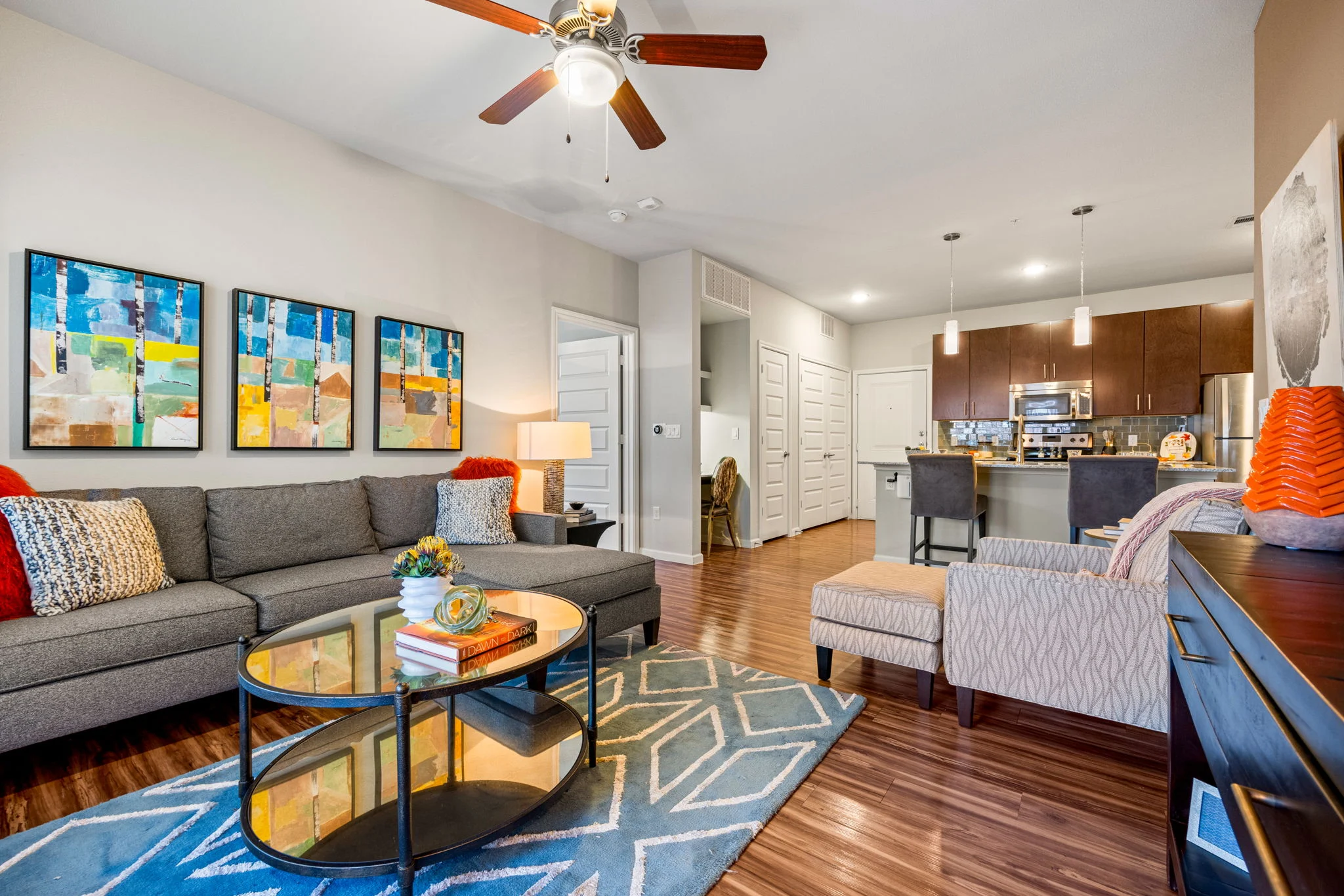 A stylish modern living room featuring a gray sofa with colorful pillows, a circular coffee table on a patterned rug, and framed abstract artwork on the wall. A kitchen is visible in the background with modern appliances and bar stools.