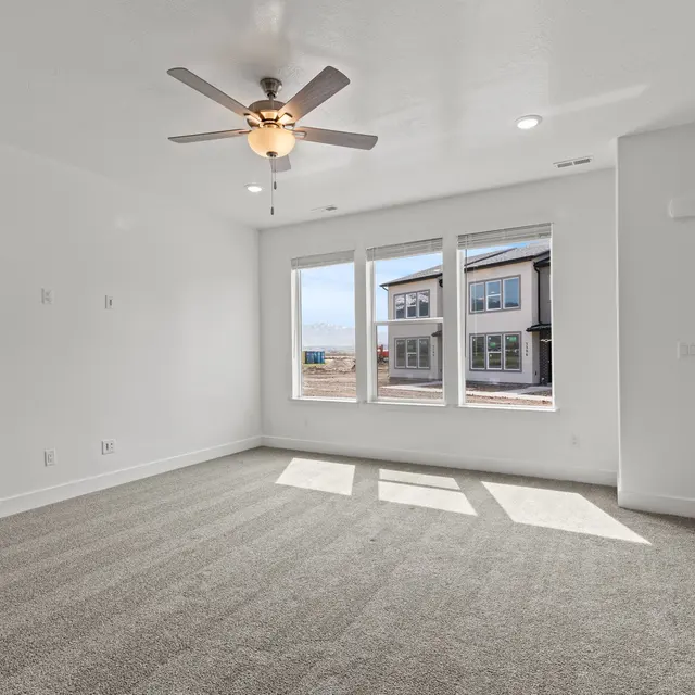 An empty living room with a ceiling fan, large windows, and a door on the right side.