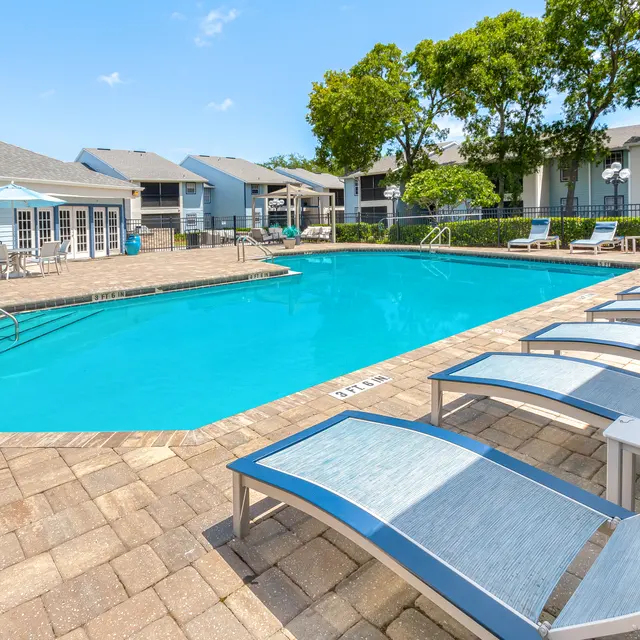 Outdoor swimming pool with lounge chairs, umbrellas, and surrounding residential buildings under a clear blue sky.
