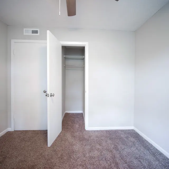 A small empty bedroom featuring light gray walls and carpet. A closet is visible with an open white door, and a ceiling fan is installed.