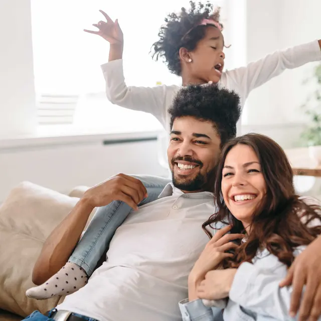 Family Joy in the Living Room A joyful family moment in a cozy living room. A man and woman smile while sitting on a sofa, with a young girl playfully posed behind them, dancing. The room has bright natural light and a plant in the background.