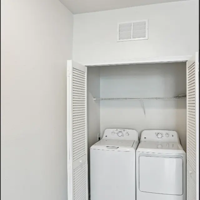 A laundry room featuring a white washer and dryer side by side, with shelves above and light-colored walls.