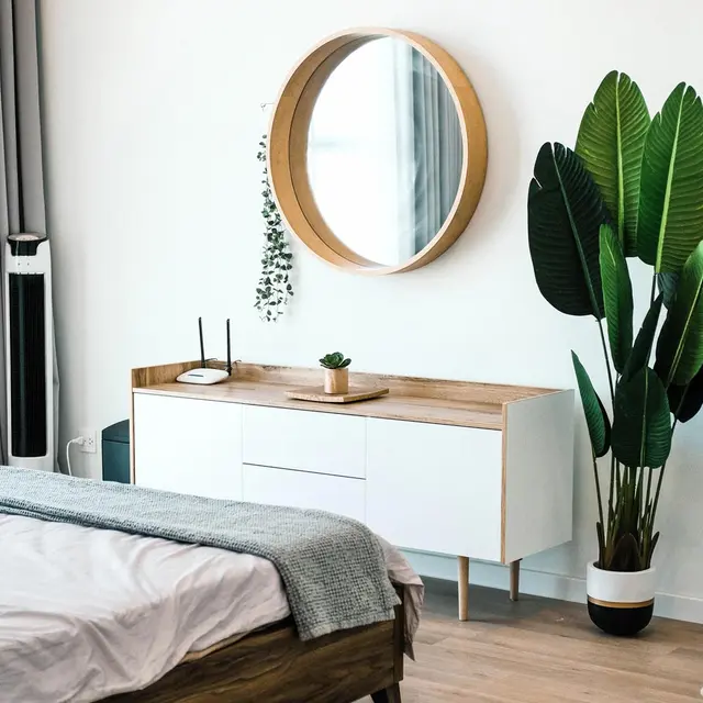 A modern bedroom with a circular mirror, a wooden dresser, and a large green plant by the wall. Light and airy decor.