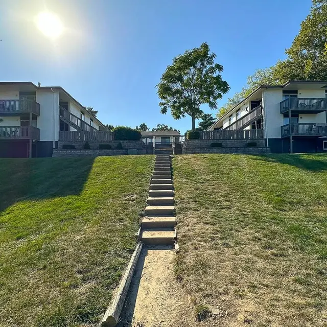 Sunny Apartment Complex Two apartment buildings on a green hill with stairs leading up to them, under a clear blue sky with the sun shining.