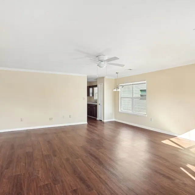 Bright and Spacious Living Room An empty living room featuring warm wood floors, light beige walls, a ceiling fan, and large windows allowing natural light to flood the space.