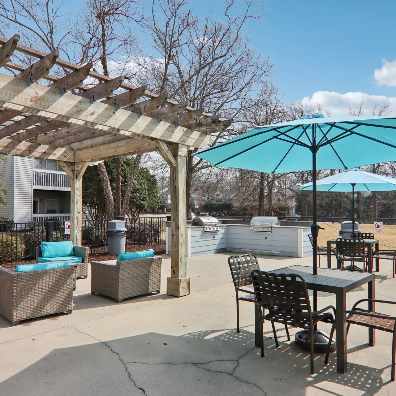 A cozy outdoor lounge area featuring a wooden pergola, blue umbrellas, and wicker furniture. There is a picnic table, additional seating, and a grill visible in the background against a partly cloudy sky.