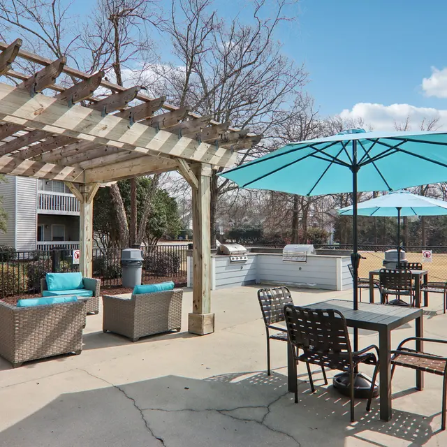 A cozy outdoor lounge area featuring a wooden pergola, blue umbrellas, and wicker furniture. There is a picnic table, additional seating, and a grill visible in the background against a partly cloudy sky.