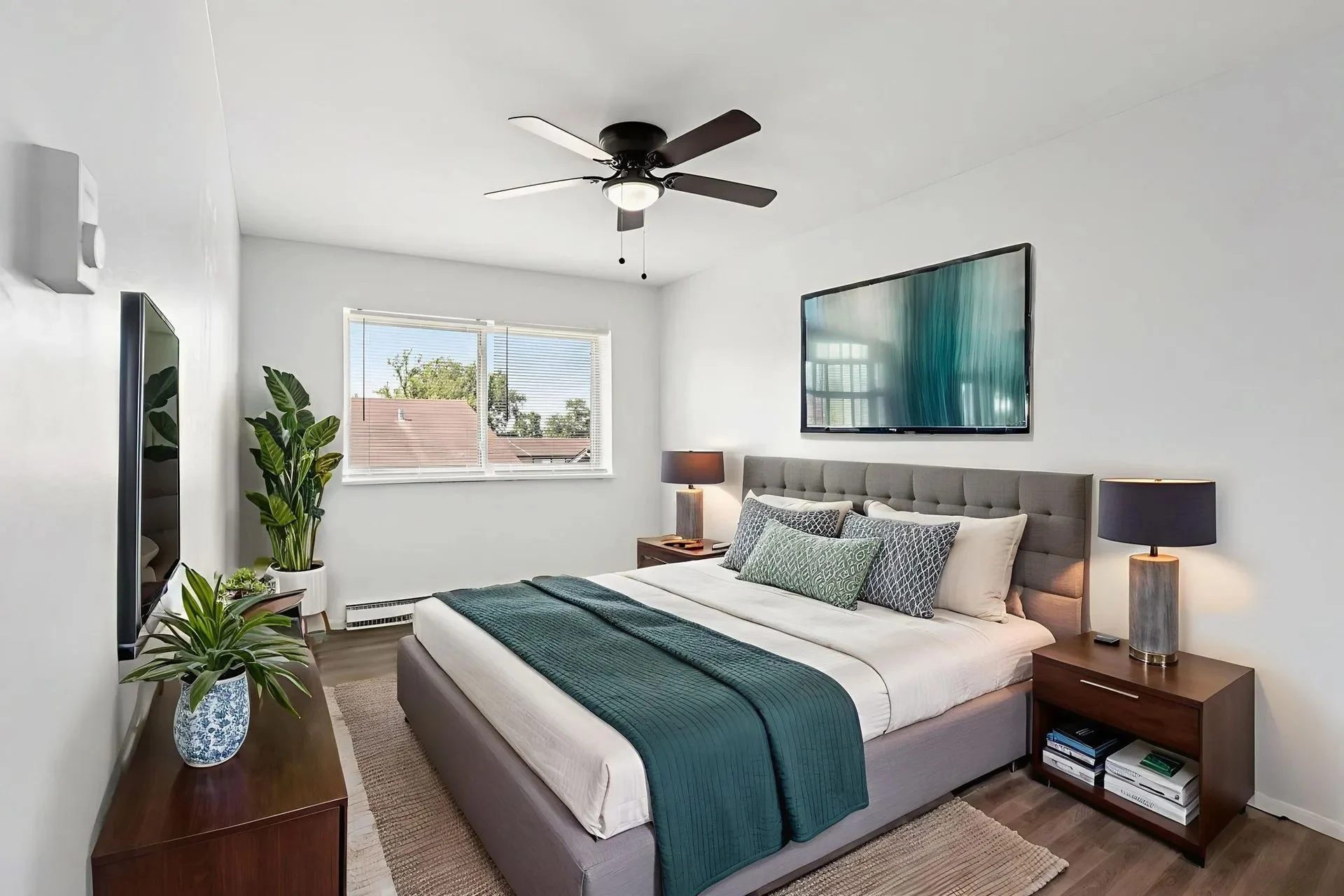 A modern bedroom featuring a large bed with a tufted headboard, decorative pillows, and a teal throw blanket. Two bedside tables with lamps flank the bed, and a large window lets in natural light. A potted plant and a wall-mounted television add to the decor.
