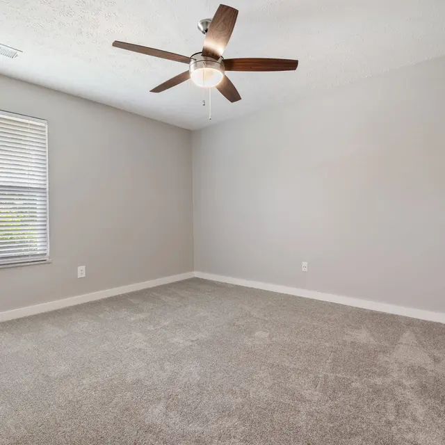 Empty room with light gray walls, a ceiling fan, and beige carpet. A window features blinds, letting in natural light.
