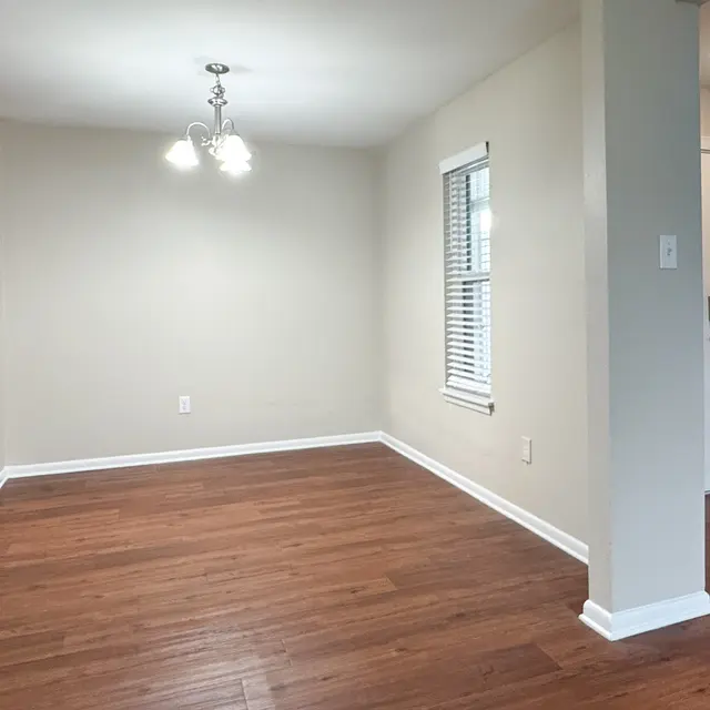 An empty living room with hardwood floors, a small chandelier, a window with blinds, and a door leading outside.