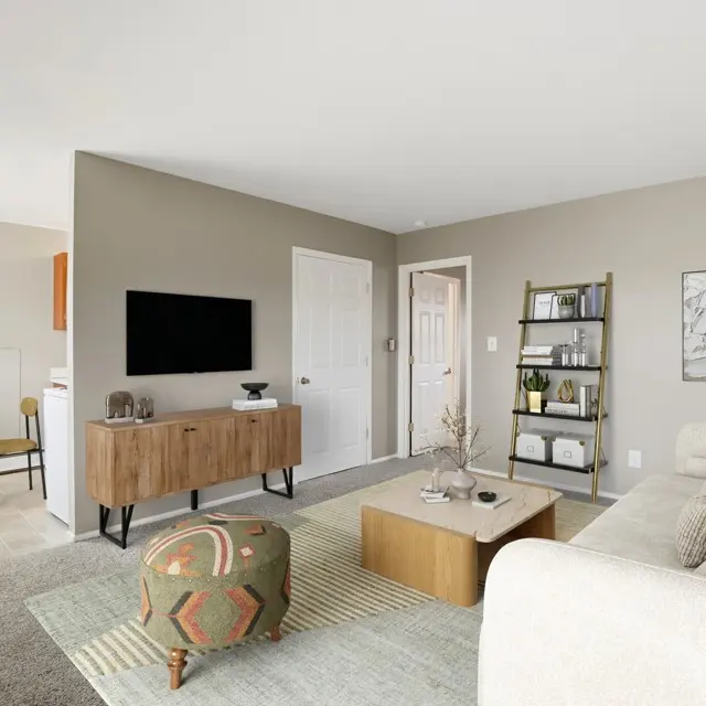 A stylish modern living room featuring a beige couch, wooden console table, flat-screen TV, and a round coffee table on a patterned rug. There is a small dining area with a table and chairs in the background, and a green decorative plant in a corner. The walls are light gray, with minimal artwork and a bookshelf.