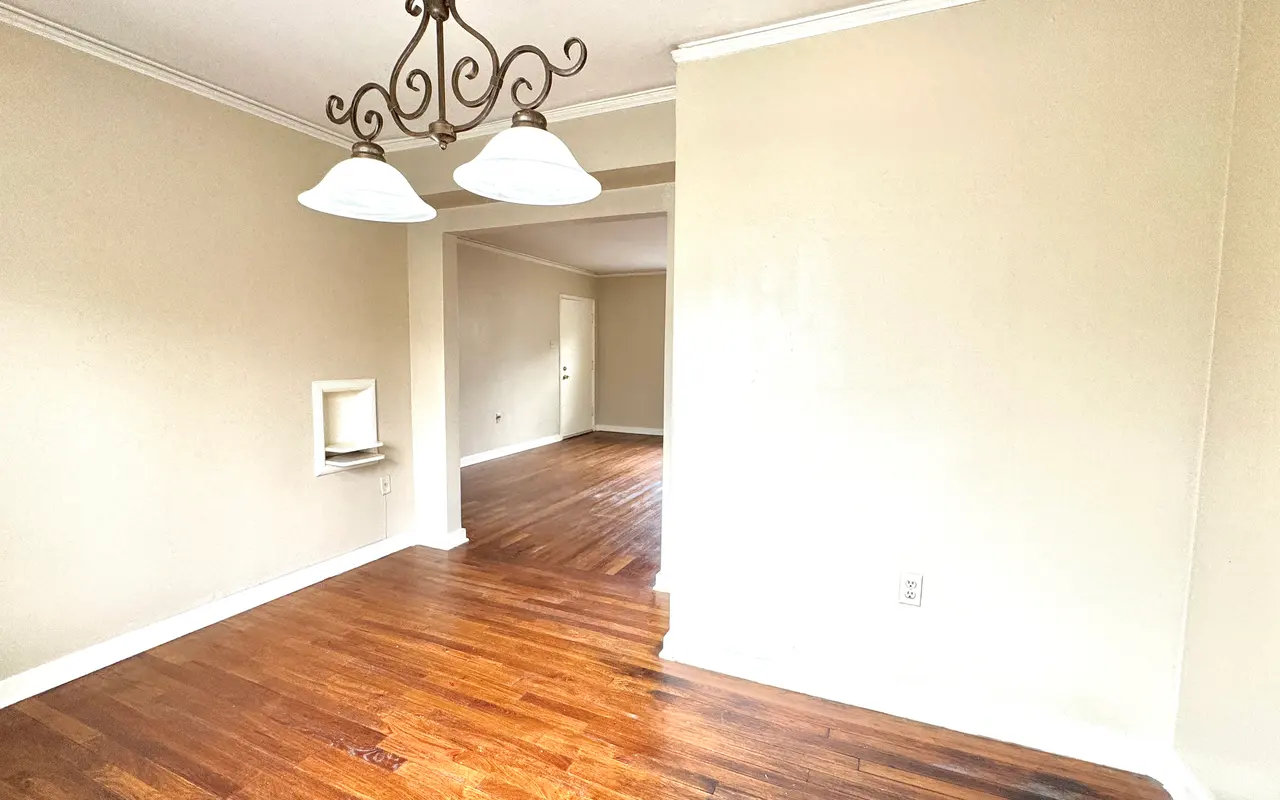 An empty dining room showing hardwood floors, beige walls, and a chandelier with two lights.
