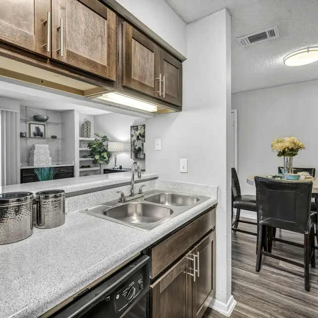 Modern kitchen with wooden cabinets, stainless steel sink, and dining area in the background featuring a round table and chairs.