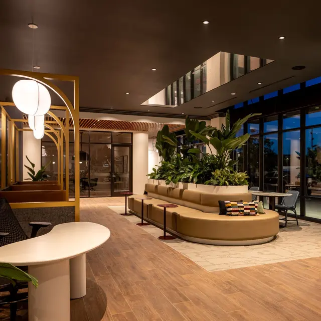 A stylish modern office interior featuring a lounge area with a curved sofa, plants, and elegant lighting fixtures. The space has large windows letting in natural light.