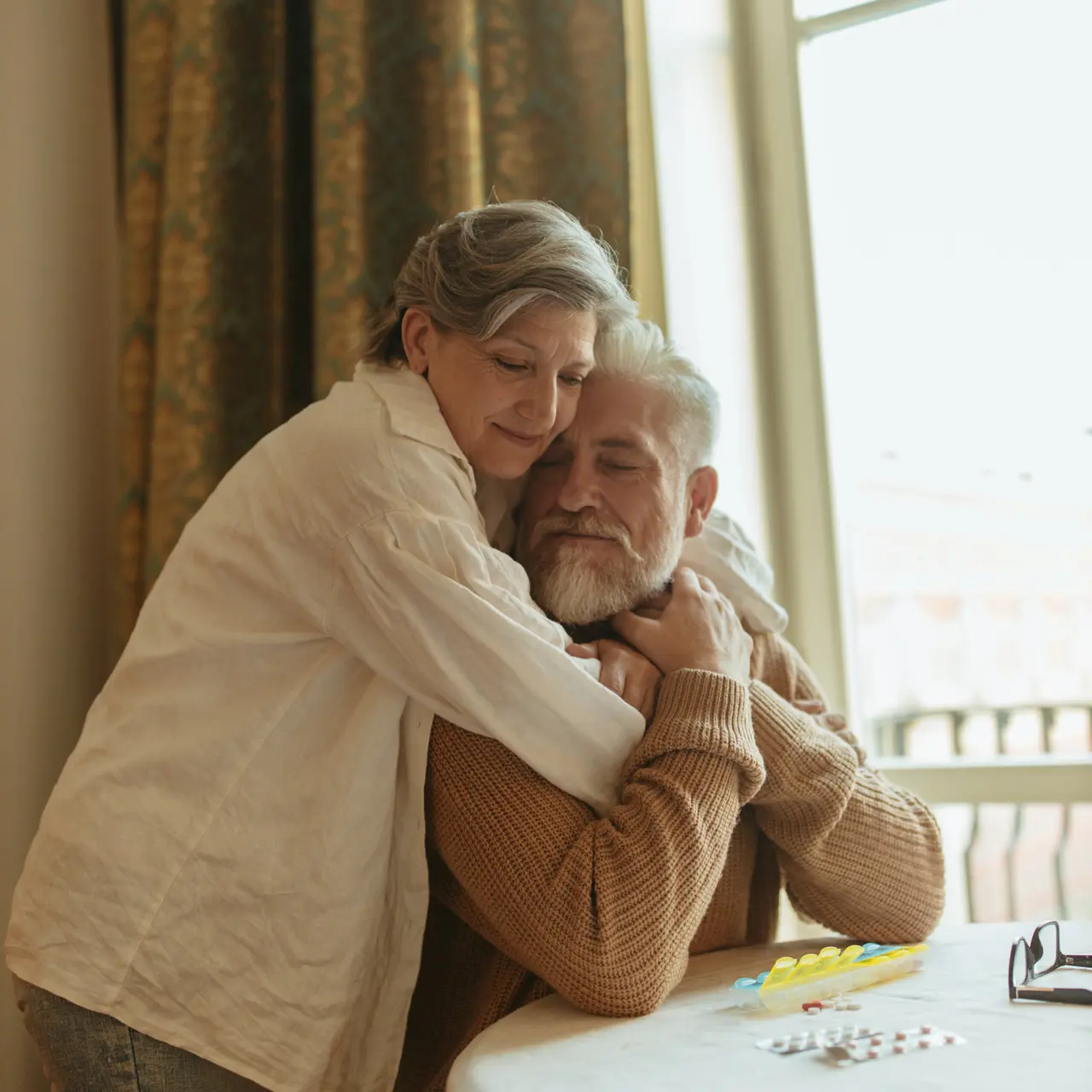 A warm embrace between an older man and woman at a table with a window in the background.