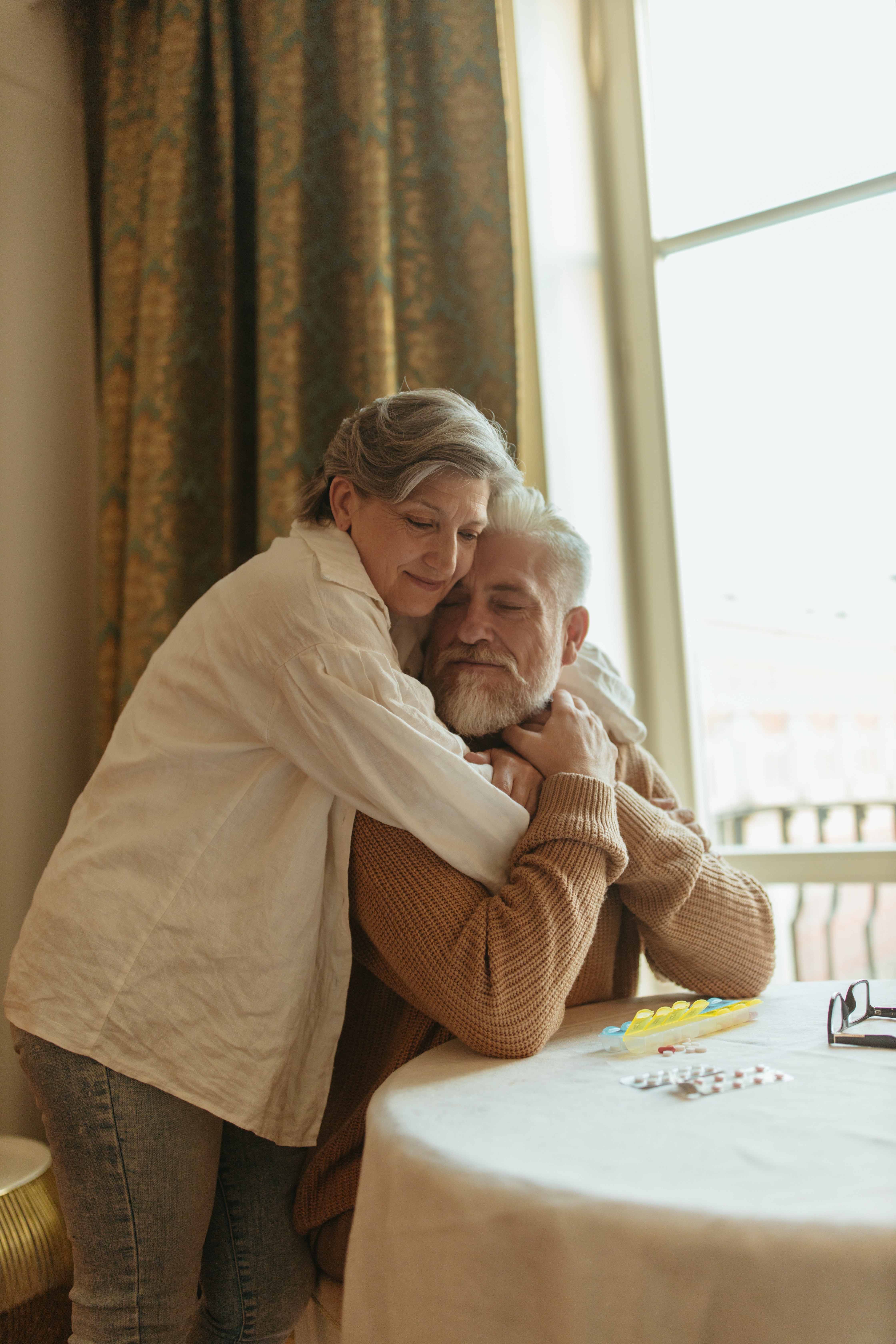 A warm embrace between an older man and woman at a table with a window in the background.