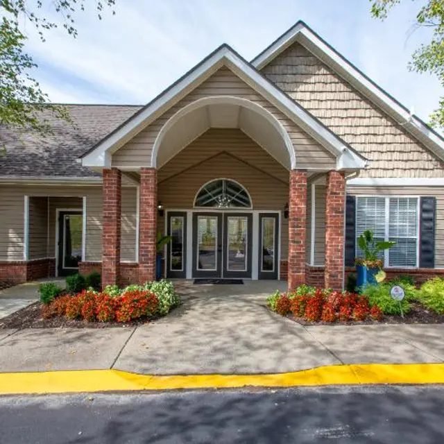 The front entrance of a community center featuring a peaked roof, large glass doors, red brick columns, and landscaping with shrubs and flowers.