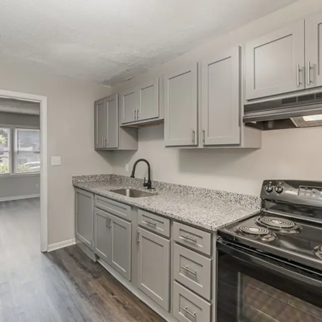 A modern kitchen featuring gray cabinets, a granite countertop, and black appliances, including a stove and refrigerator. The kitchen is spacious and connects to a room with large windows.