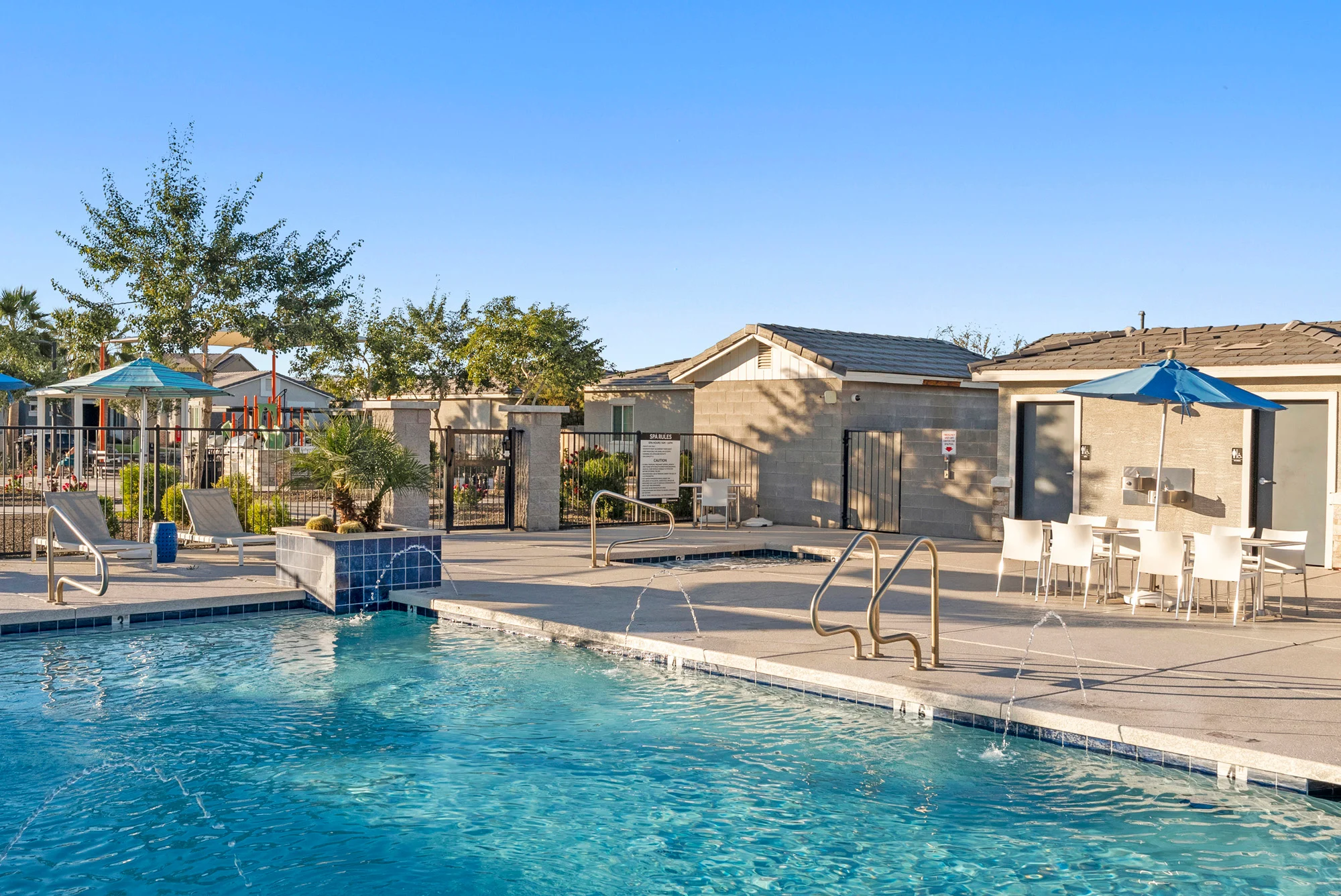 A clear blue swimming pool with a fountain feature, surrounded by lounge chairs and patio umbrellas. In the background, there are buildings and trees. The sun is shining brightly in a clear sky.
