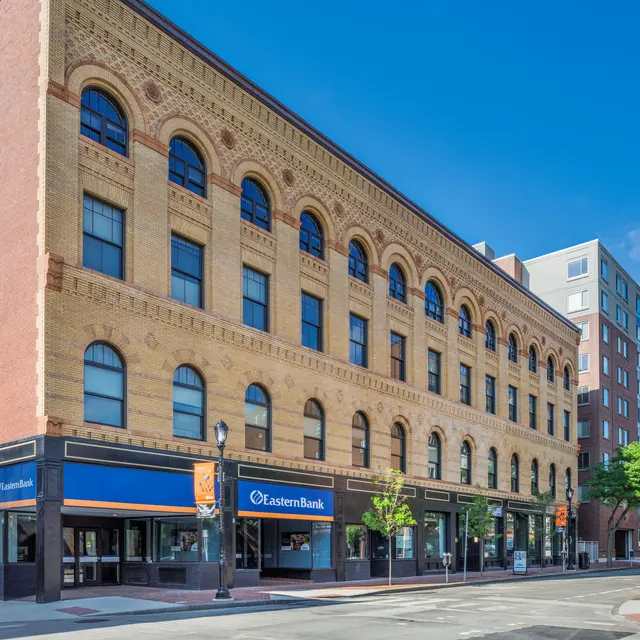 A historic brick building with arched windows featuring the signage of Eastern Bank. The structure has four floors with detailed architectural elements and is situated at a street corner, surrounded by modern buildings and greenery.