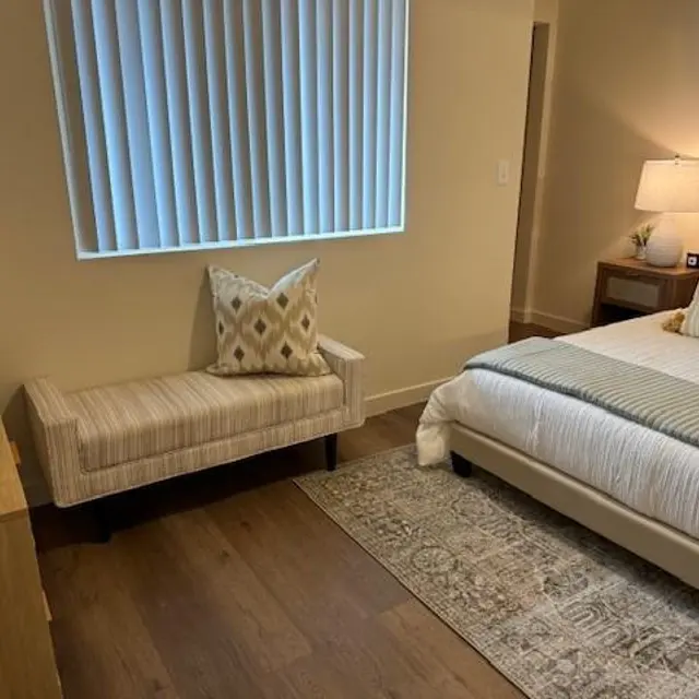 A cozy modern bedroom featuring a bed with a patterned quilt and pillows, a decorative bench with a cushion, and a bedside table with a lamp. There are wooden floors and a window with vertical blinds.
