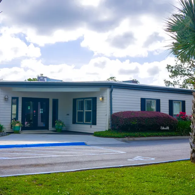 A single-story building with a modern design featuring large windows, surrounded by well-maintained landscaping including palm trees and shrubs with flowers.