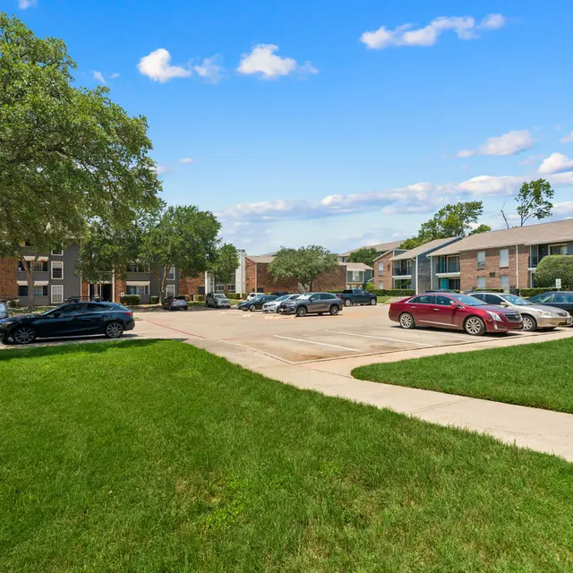 An outdoor view of an apartment complex parking lot surrounded by grass and trees, showcasing several parked cars and blue sky.