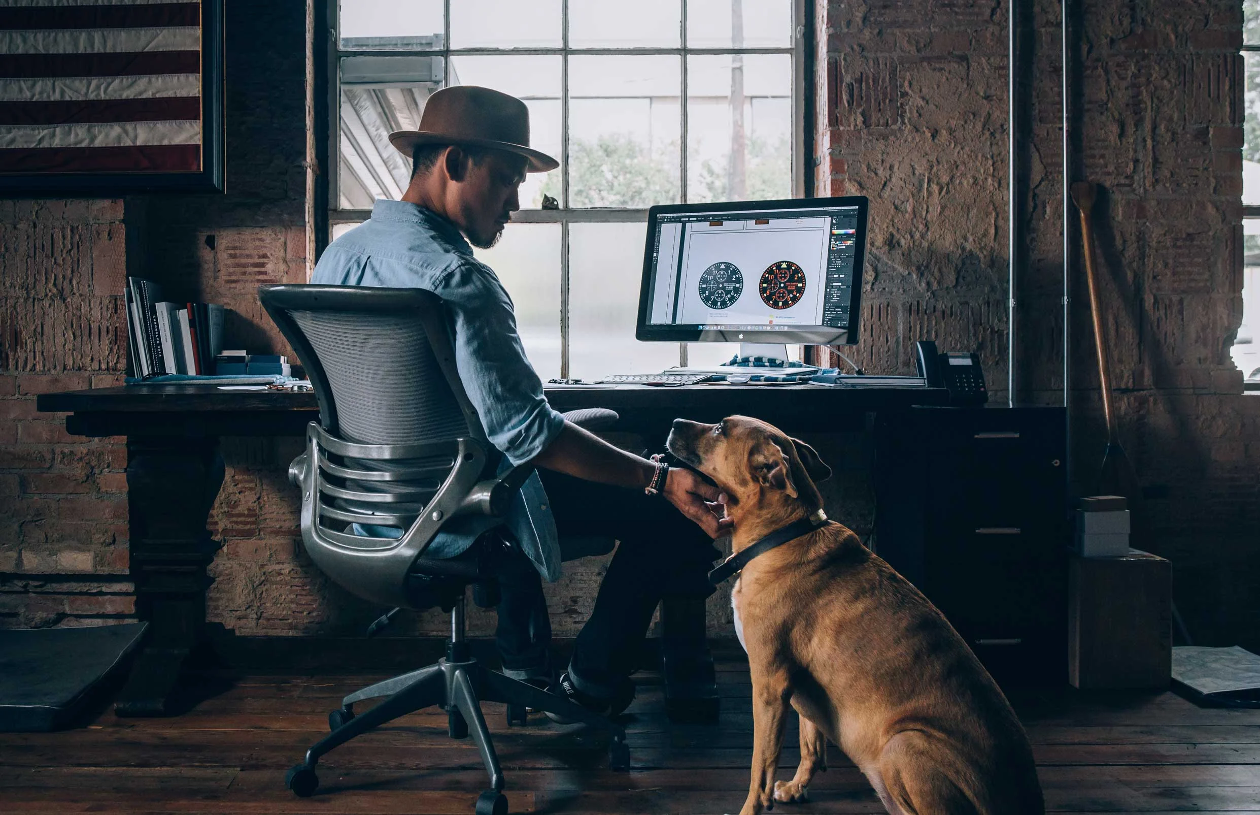 A person sitting at a desk in an office, petting a dog. The room has a rustic feel with wooden floors and a large window letting in natural light. A computer monitor displays colorful designs on the desk.