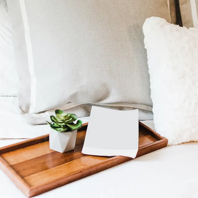 A cozy bedroom setup featuring a wooden tray with a succulent plant and a piece of blank white paper, resting on a bed with decorative pillows.