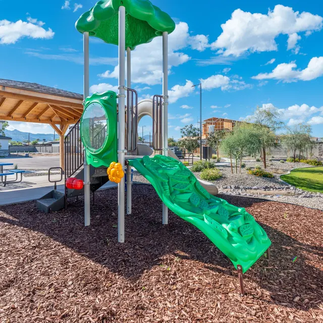 A colorful playground featuring a green slide and climbing structure, surrounded by mulch and grass, with a picnic area in the background.