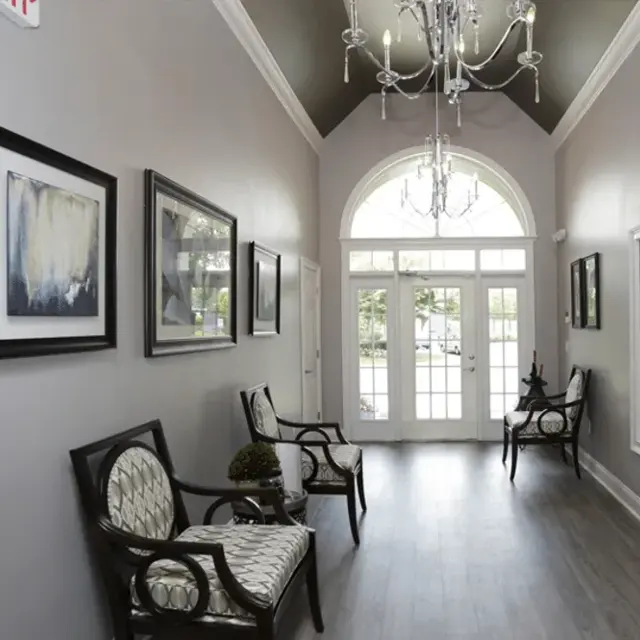 A stylish hallway featuring two chairs, decorative artwork on the walls, a chandelier, and large windows allowing natural light in. The floor is wooden, and the overall decor is modern and elegant.