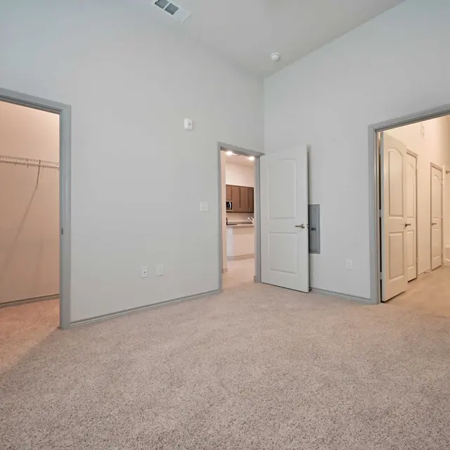 A spacious room with light gray walls and carpet. Two doors lead to separate closets or rooms, one visible on the left and the other on the right, with a clear view of a kitchen area through an open door in the background.