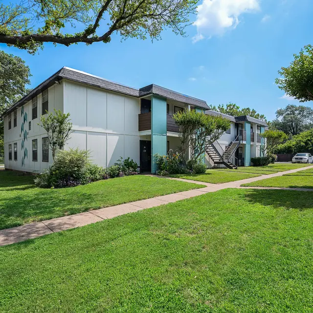 Exterior view of a two-story apartment complex surrounded by green grass and trees on a sunny day.