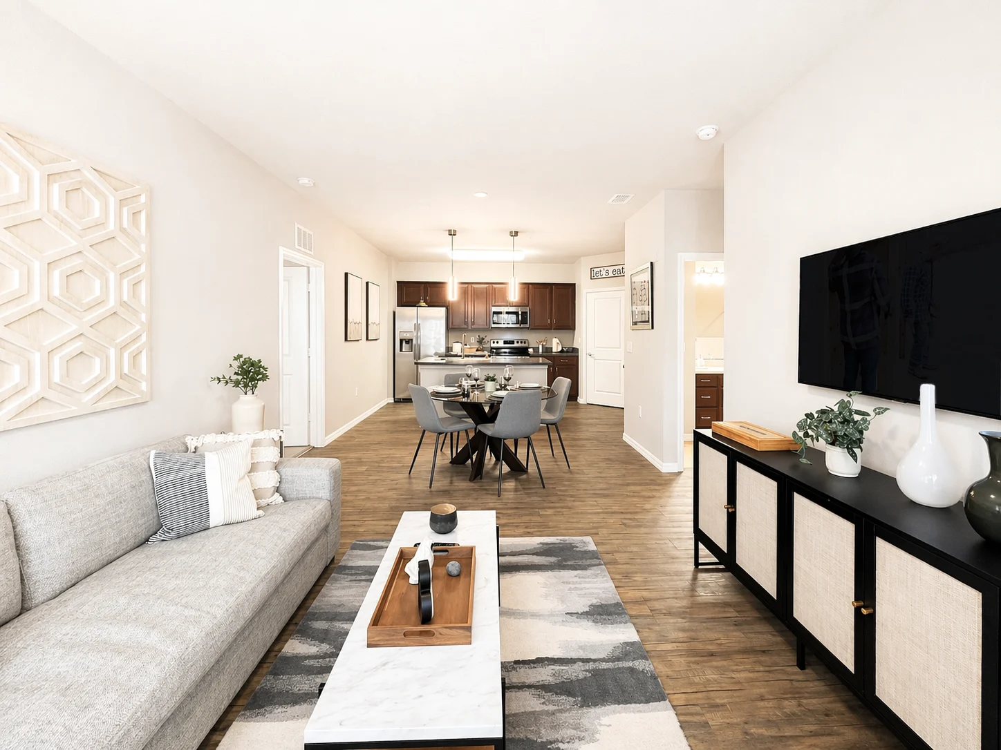 A modern living room featuring a gray sofa, a coffee table with decorative items, a dining area in the background, and a black television stand with plants and vases.