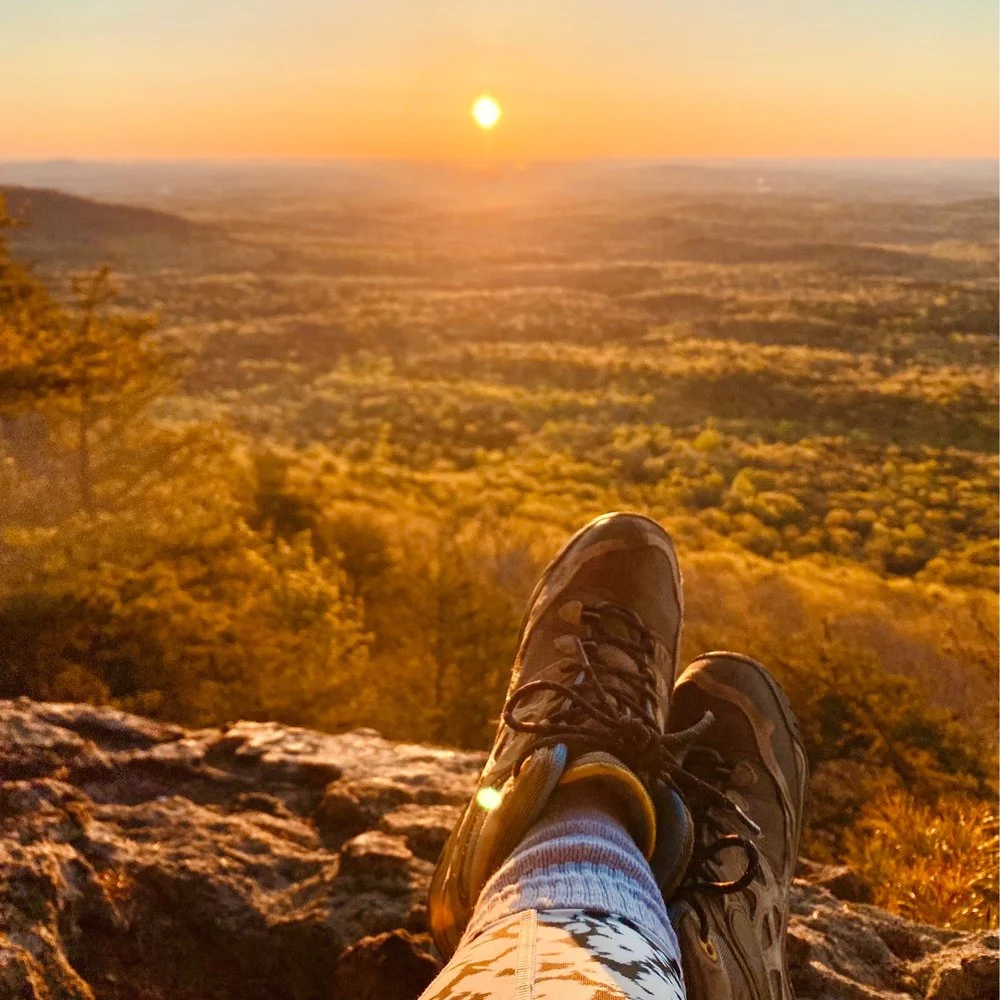 Serene Sunset from a Hiking Spot A person sitting on a rocky outcrop wearing hiking shoes and patterned leggings, with feet resting on the rocks, overlooking a picturesque sunset landscape with hills and valleys creating a scenic view.