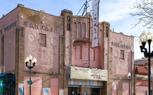 An exterior view of the Santa Fe Theatre, showcasing its pink facade with faded signage and lights. The building features decorative elements and is partly covered with street art. Nearby street lamps are visible in the foreground.
