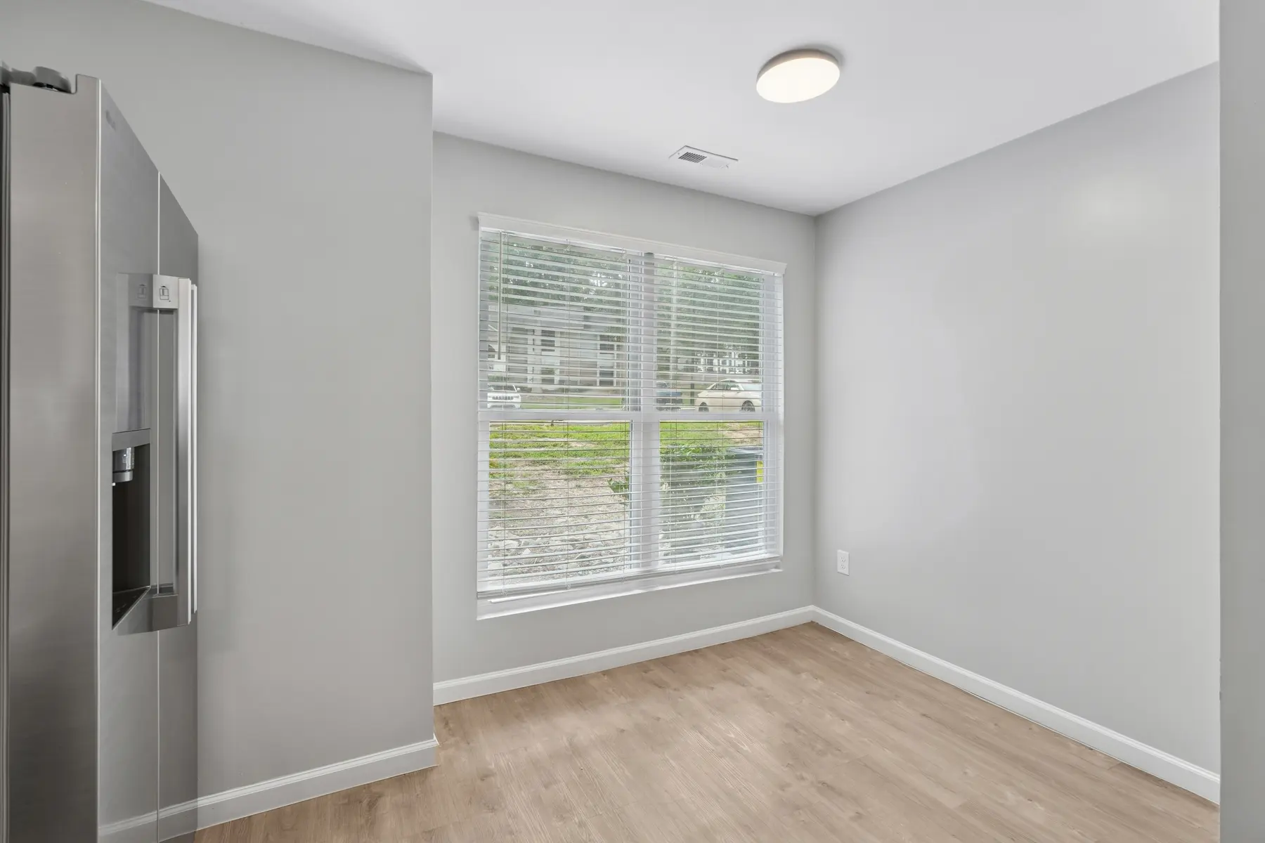 A modern kitchen corner featuring a window with blinds, pale gray walls, and hardwood flooring. A stainless steel refrigerator is visible on the left side.