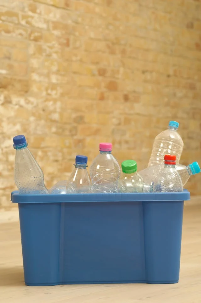 Recycling Plastic Bottles in a Blue Bin A blue recycling bin filled with various plastic bottles, some capped with colorful lids. The background features a textured brick wall, enhancing the focus on the bin.