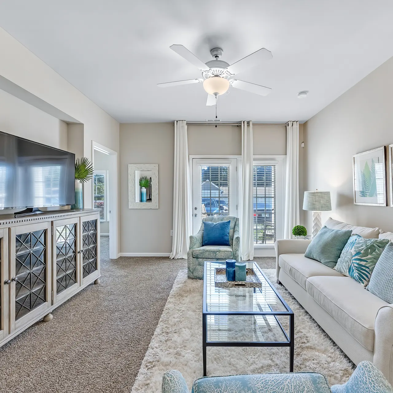 A modern living room featuring a comfortable sofa, decorative pillows, a coffee table, and a decorative cabinet with a TV. Light colored walls and large windows provide natural light.