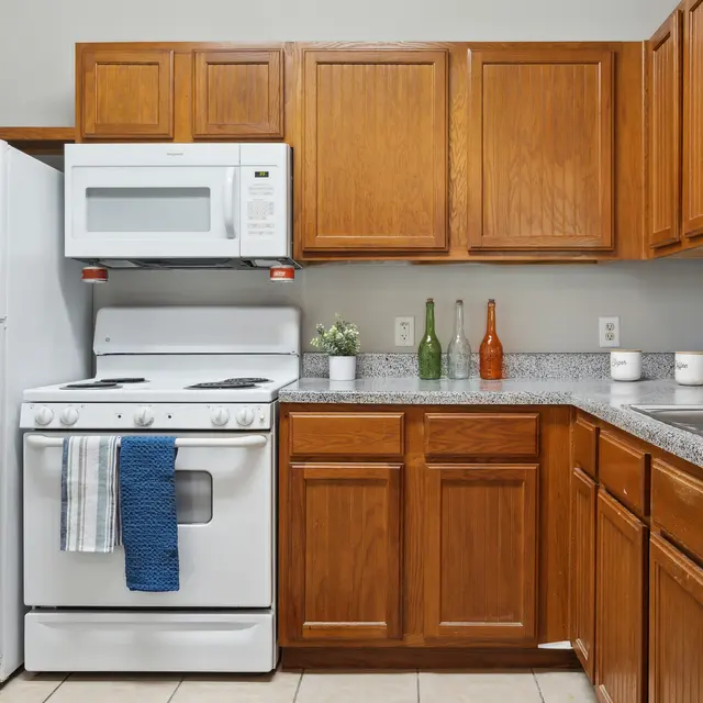 A cozy kitchen with wooden cabinets, a white stove, microwave, and refrigerator, featuring a gray countertop and decorative bottles.