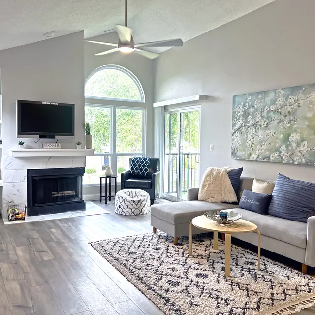 A modern living room featuring a gray sofa, decorative pillows, a small round coffee table, and a patterned rug. There's a large window allowing natural light, a TV mounted above a fireplace, and decorative shelves with items. A chair and ottoman are in the corner, and the walls are painted in soft tones with artwork hanging.
