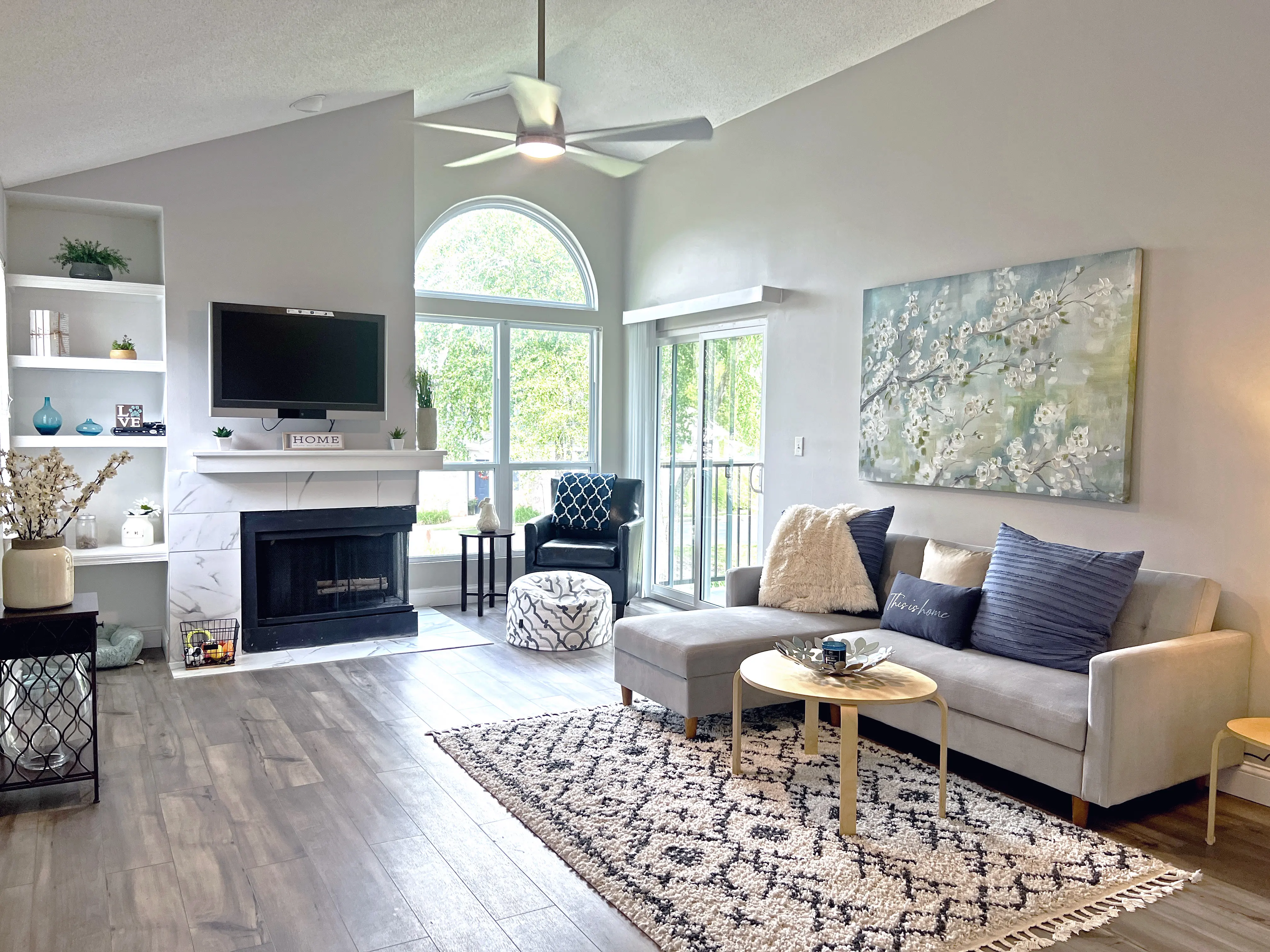 A modern living room featuring a gray sofa, decorative pillows, a small round coffee table, and a patterned rug. There's a large window allowing natural light, a TV mounted above a fireplace, and decorative shelves with items. A chair and ottoman are in the corner, and the walls are painted in soft tones with artwork hanging.