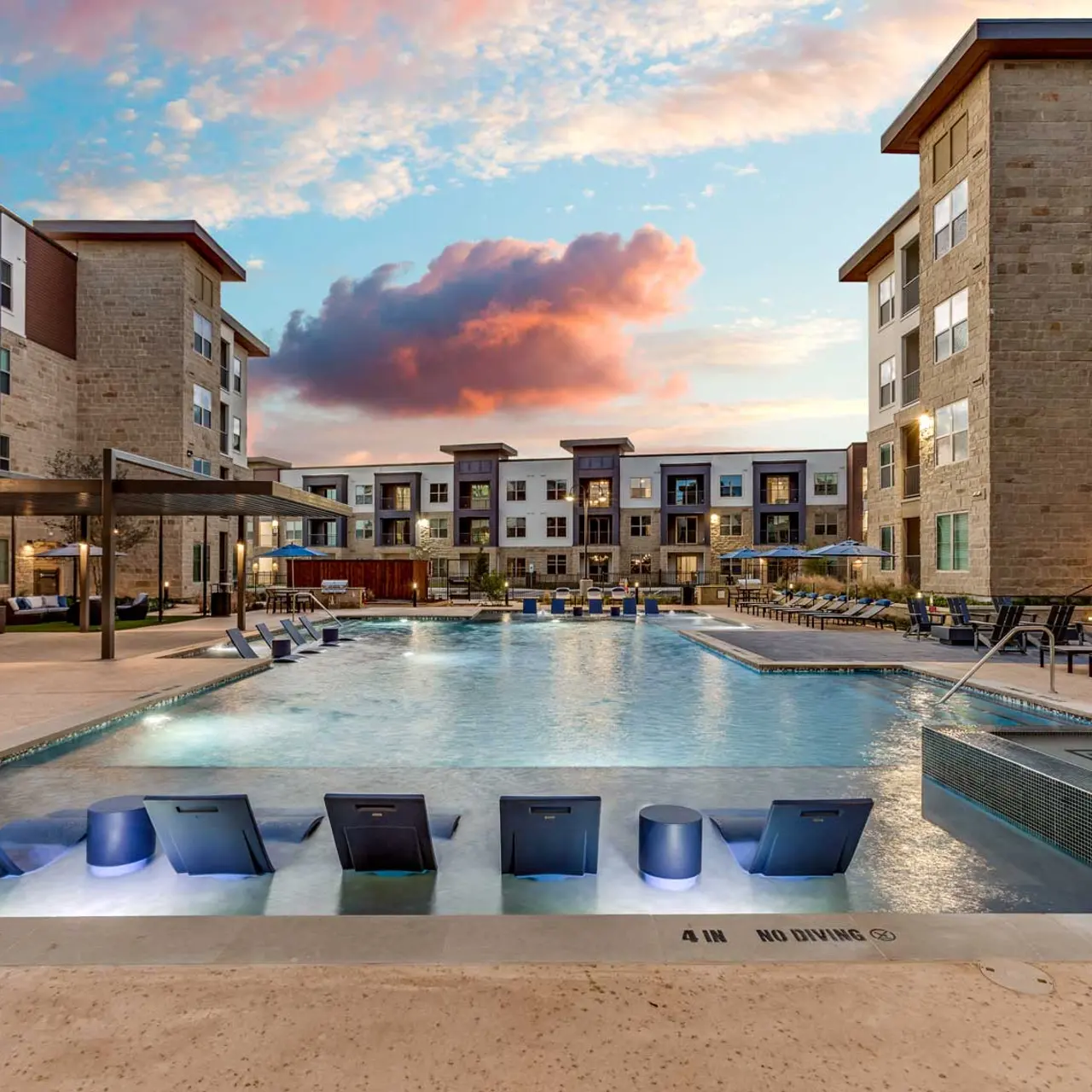 Modern Apartment Complex Pool A modern apartment complex pool area featuring a large swimming pool with loungers, surrounded by buildings at sunset.