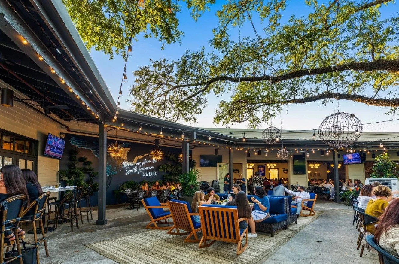 A vibrant outdoor dining area filled with people, featuring blue lounge chairs, string lights, and a lively atmosphere under trees.