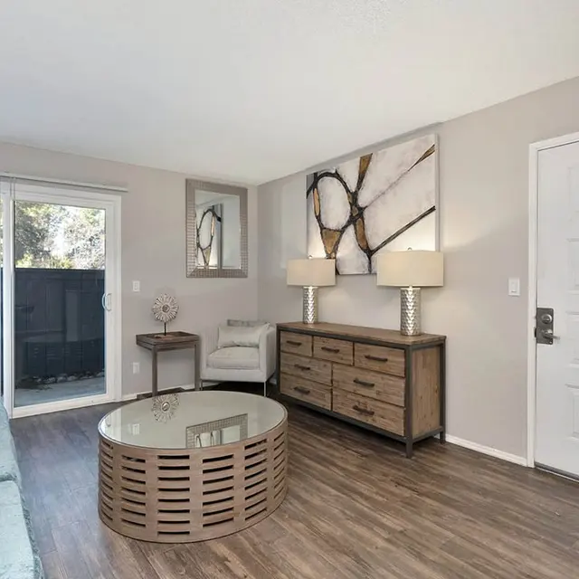A modern living room featuring a light blue couch, a round coffee table, two lamps on a wooden dresser, and a sliding glass door leading to an outdoor area.