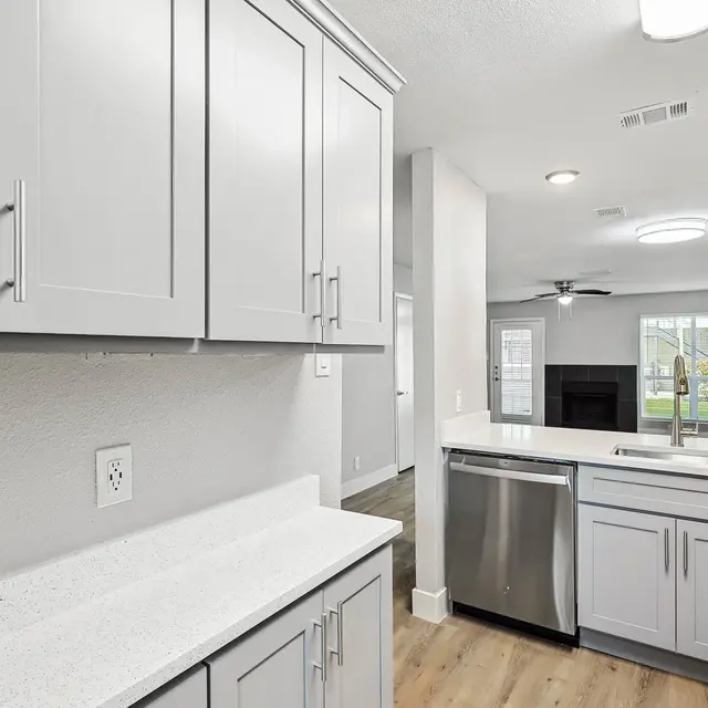 A modern kitchen featuring light gray cabinets, a white countertop, stainless steel appliances, and a spacious layout leading to a living area.